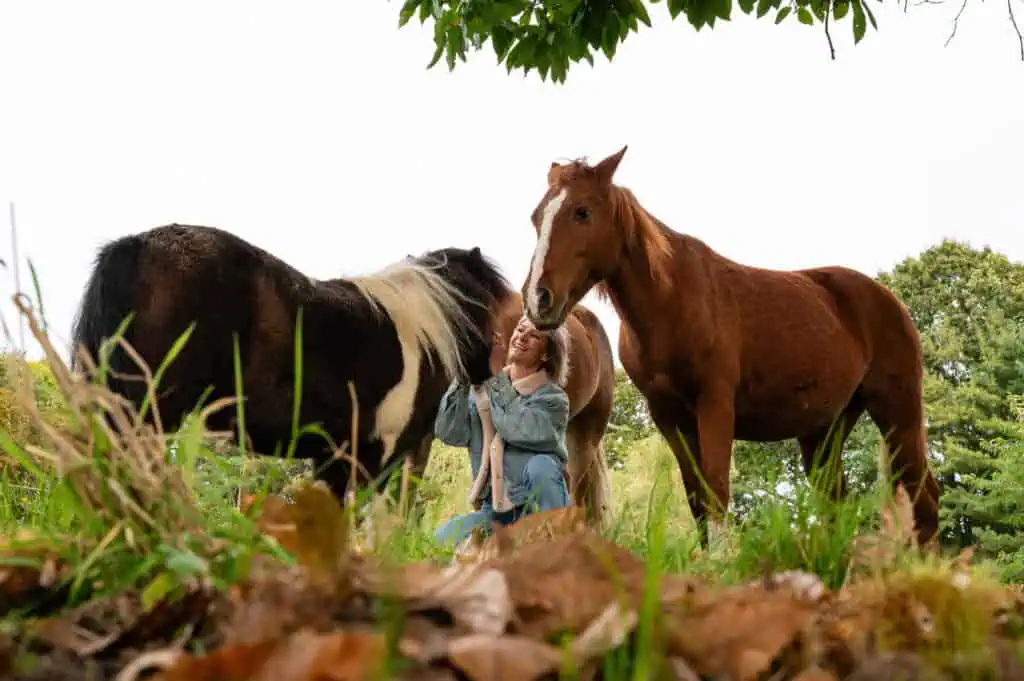 Alt tekst: Vrouw knuffelt paarden in weide, groene omgeving, gelukkig, ontspannen, puur genieten, omringd door natuur en dieren, perfecte moment voor paardenliefhebbers.