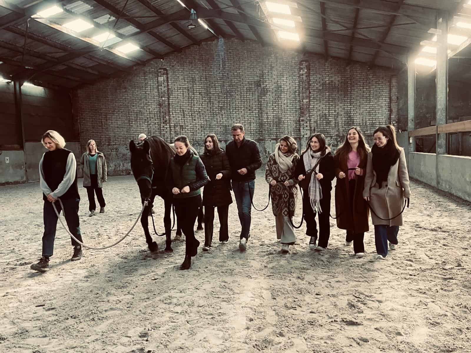 Cadeaus Strook, stijlvol wandelend op een paard in een paardenstal, groep vrouwen gezellige dag, educatieve activiteit, teamuitje, paardrijden in een binnenbak, heren en dames, casual en comfortabele kleding, sfeer van vriendschap en ontspanning, les of training in paardensport, veiligheid en plezier, informele groepsactiviteit, mooie omgeving, brainstormsessie of workshop, plezier bezig met dieren, fotograferen en leren, plezierige dag, bedrijfsevenement, ontspannend uitje voor vriendinnen, team building, passie voor paarden, agrarische omgeving, ontspanning en plezier, leren en genieten, pedagogische activiteit, teamuitje met dieren, voorliefde voor paarden, samen lachen en connectie maken, buitenactiviteiten, fotografie van een mooie dag, belevenis in een paardenpiste, informele groepssfeer.